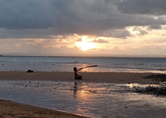 Didjeridoo Musicien assis sur le sable qui joue du didgeridoo face à l’océan, avec un magnifique coucher de soleil à l’horizon et un ciel nuageux en arrière-plan