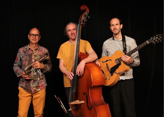 Dume Trois musiciens posant avec leurs instruments devant un fond noir : un trompettiste portant une chemise colorée et un pantalon jaune, un contrebassiste en t-shirt ocre et un guitariste vêtu d’une chemise blanche à motifs, tenant une guitare électrique jazz