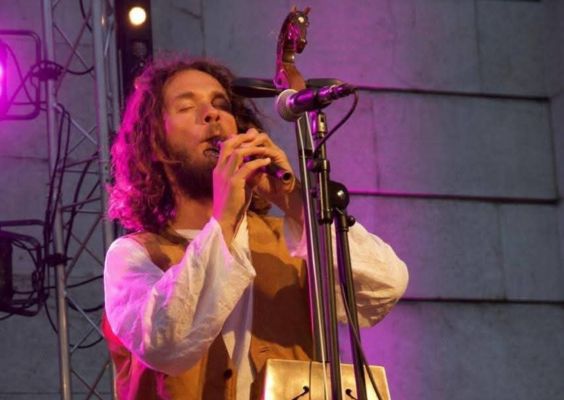 Instruments-exotiques Musicien aux cheveux longs, vêtu d’un gilet en cuir et d’une chemise blanche, jouant d’un instrument à vent traditionnel sur une scène éclairée de lumières violettes et oranges