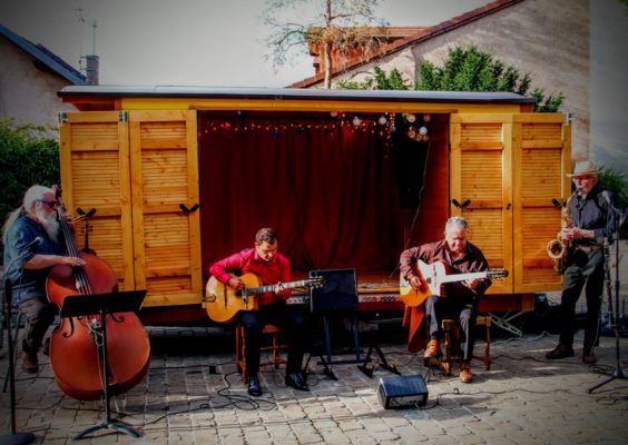 Le-Chemin-des-Nuages Un quartet de musiciens joue devant une petite scène mobile en bois : un contrebassiste barbu, deux guitaristes assis et un saxophoniste debout, dans une ambiance chaleureuse et intimiste