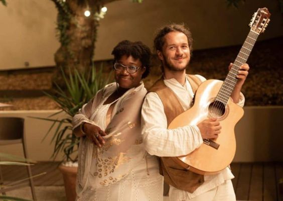 Sunshine-Melody Duo de musiciens qui pose avec le sourire : une chanteuse en robe blanche ornée de détails dorés et un guitariste vêtu d’un gilet beige et d’une chemise blanche, dans une ambiance chaleureuse de jardin éclairé