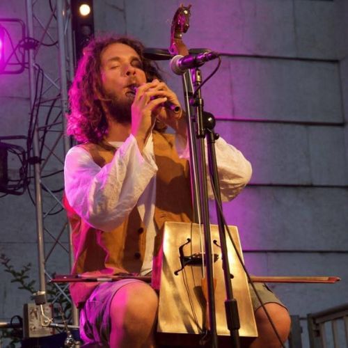 Flo-del-mundo Musicien aux cheveux longs, vêtu d’un gilet en cuir et d’une chemise blanche, jouant d’un instrument à vent traditionnel sur une scène éclairée de lumières violettes et oranges