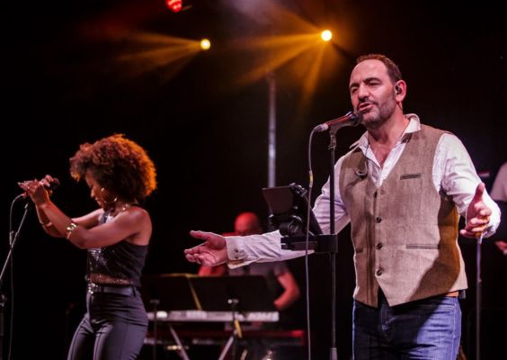 Groupe de six musiciens jouant sur une scène sous des projecteurs colorés. Trompette, clarinette, contrebasse et banjo accompagnent une chanteuse et un batteur dans une ambiance jazz/folk
