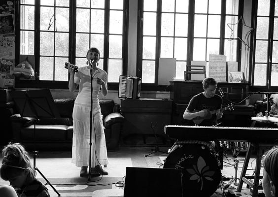 Duo-sequoia Duo en concert dans une salle lumineuse : une chanteuse en robe blanche au micro avec un tambourin, accompagnée d’un musicien jouant du ukulélé, entouré d’instruments